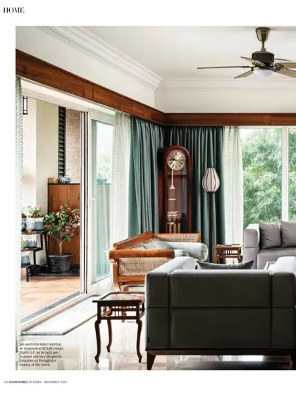 The living room connects seamlessly to a double-height deck, blurring the lines between indoors and outdoors. The design balances modern grey sofas with antique wooden furniture, like the stately grandfather clock.
