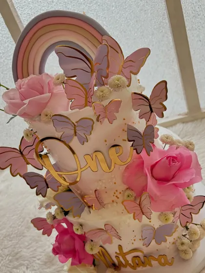 A stunning butterfly-themed cake for a first birthday. This two-tier cake features a pastel rainbow, fresh roses, and a cascade of pink and purple butterflies.