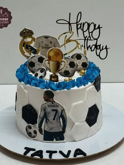A football-themed cake featuring Cristiano Ronaldo. The sides of the cake are decorated with a soccer ball pattern, and the top includes a trophy and other football-related toppers.