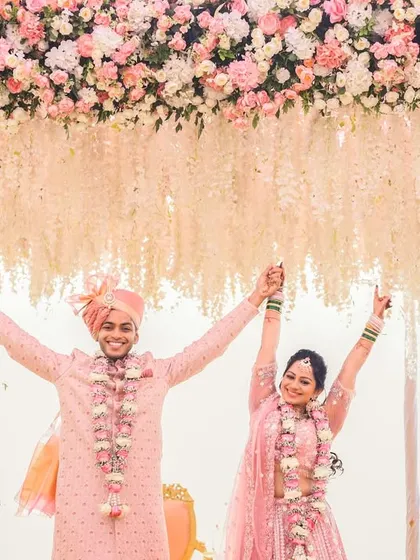A moment of pure triumph and joy under the floral mandap. The couple raises their hands in celebration after completing their wedding rituals, perfectly framed by the beautiful pink and white decor.