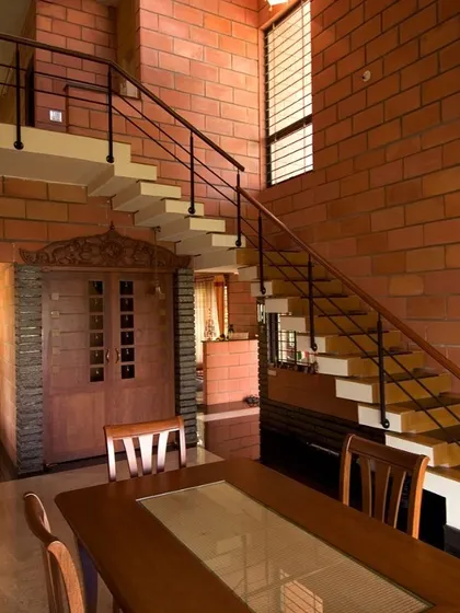 At the Sharada Residence, an exposed brick wall provides a dramatic backdrop for the dining area and floating staircase. The design integrates a traditionally carved wooden door, blending rustic materials with classic Indian craftsmanship.