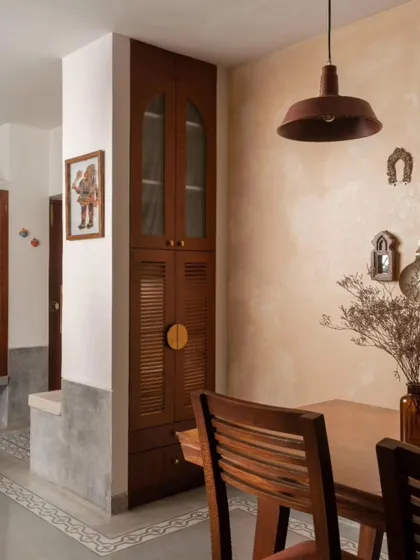 A view of the dining space, showing the simple wooden dining set and the industrial-style pendant light, which contrast with the traditional form of the storage unit.