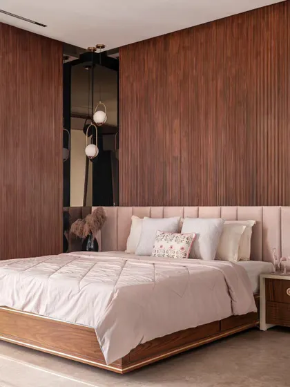 A close-up of a bedroom designed with warm, natural materials. The vertically paneled wood wall adds texture and depth behind the bed. The soft pink bedding and headboard create a gentle and inviting feel.
