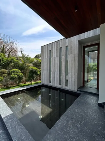 At the entrance of Rosewood Farmhouse, a shallow water feature adds a sense of tranquility and elegance. The dark water reflects the sky and the warm wood ceiling of the porch, creating a peaceful welcome that aligns with Vastu principles.