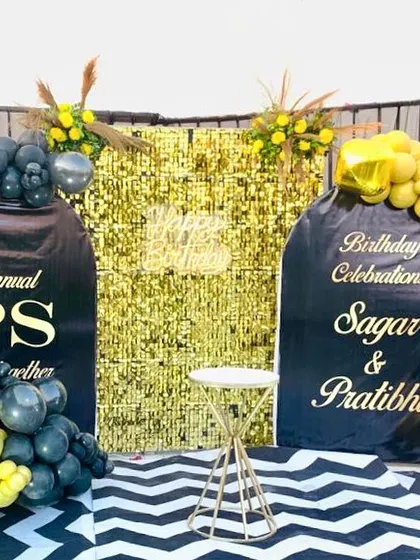 A custom birthday celebration for a couple. The setup includes a gold shimmer wall and personalized backdrops with black and yellow balloon arrangements.