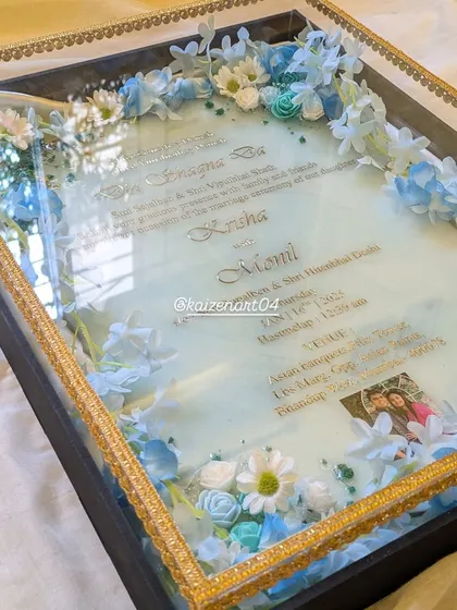 A detailed shot of a custom wedding Kankotri in a beautiful blue and white theme. The intricate details of the text and the delicate arrangement of flowers are all sealed under a glossy resin finish.