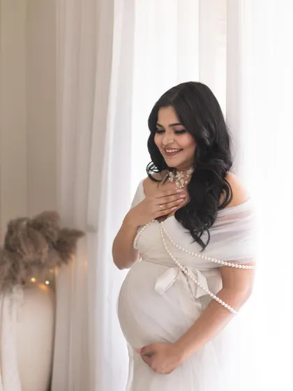 A candid, smiling portrait of the mother-to-be. The soft, natural light from the window and the delicate pearl accessories add to the angelic and serene feel of this shot.