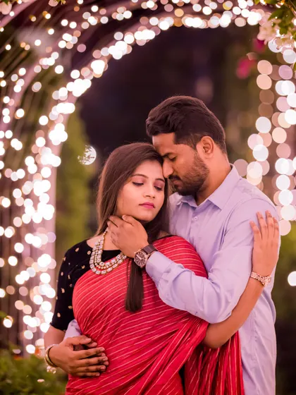 A beautiful, intimate portrait of a couple in a saree and modern attire, framed by a tunnel of lights. The focus is on their close bond and shared emotion.