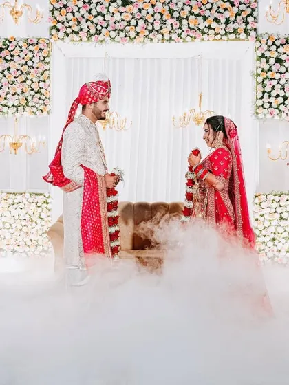A dreamy Varmala moment on a stage surrounded by low-lying fog, with a backdrop of white florals and chandeliers.
