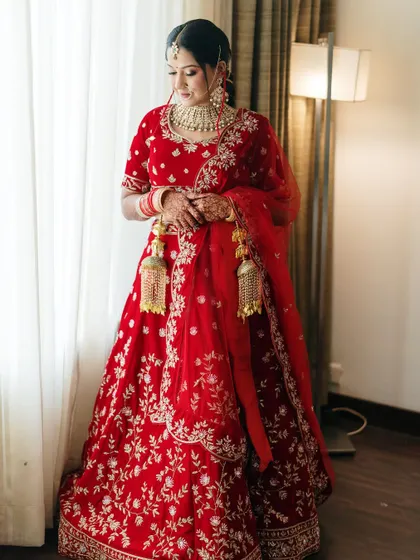 A full-length bridal portrait by the window. The soft, diffused light creates a classic and elegant look, highlighting the rich red of her lehenga.