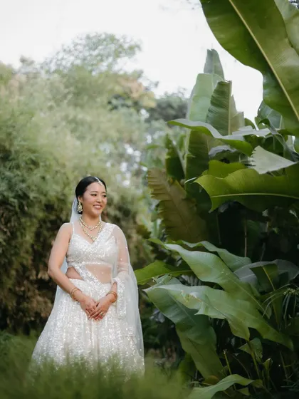 A bride in a shimmering white lehenga stands amidst lush greenery, her smile radiating happiness and serenity.