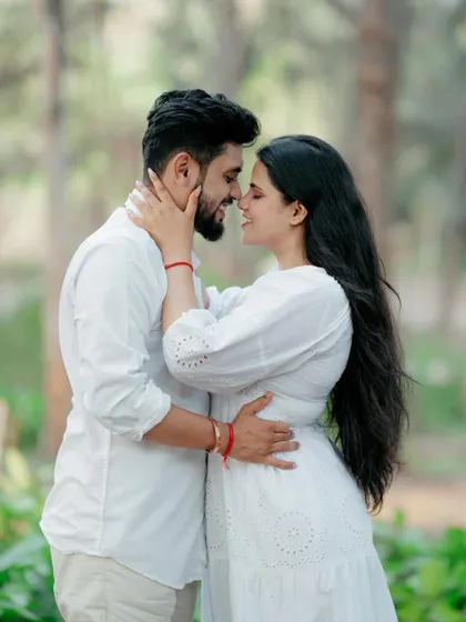A classic romantic portrait of a couple in a forest, standing close and touching foreheads. The lush green background makes their white outfits stand out beautifully.