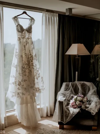 An artistic shot of the bride's wedding dress hanging by a window, with her veil and bouquet resting on a chair.