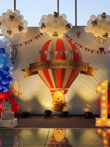 A grand first birthday celebration with a hot air balloon theme. This custom backdrop features a large balloon with a teddy bear, surrounded by balloon clouds and a lit-up "ONE" marquee sign, creating a perfect photo opportunity.