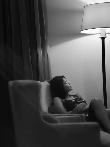 A quiet, contemplative black and white portrait of a woman sitting in an armchair, illuminated by a single floor lamp. The deep shadows and focused light create an intimate and moody atmosphere.