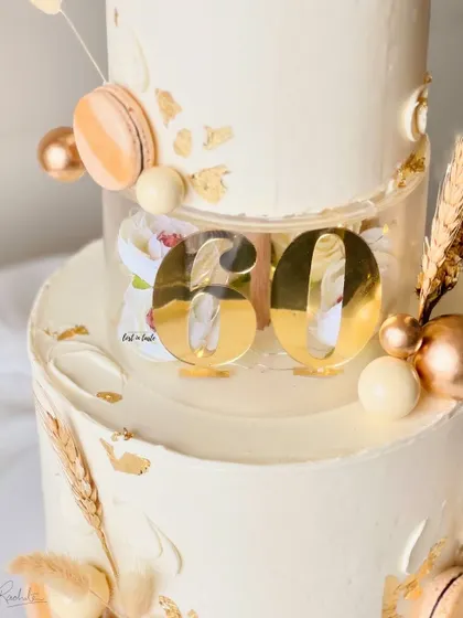 A sophisticated two-tier cake for a 60th birthday celebration in an ivory and gold theme. This design features a clear separator tier, macarons, and dried palm leaves for a modern, celebratory feel.