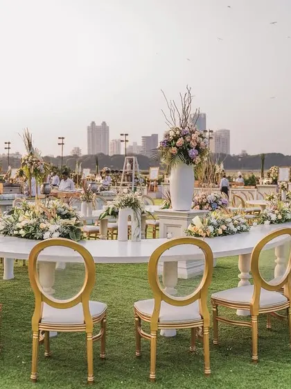 An elegant, serpentine dining table set for guests on the lush green lawn. The white table, paired with gold-accented chairs and fresh floral runners, provided a sophisticated dining experience with a view of the entire celebration.
