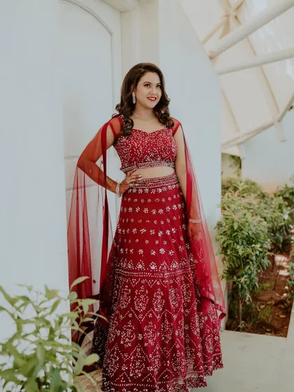 A full-length standing shot of the bride-to-be. Her red lehenga is paired with sheer cape-style sleeves, adding a modern touch to her engagement outfit.