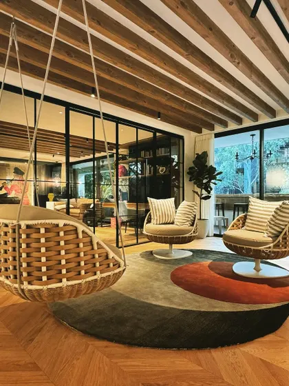 We created this informal lounge area as a peaceful escape from the formal settings of the house. A woven hanging swing chair and comfortable swivel chairs are arranged on a circular rug, encouraging relaxation and conversation. The space is defined by its connection to the outdoors and its biophilic design elements, making it a perfect spot to unwind.