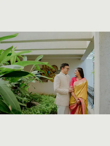 A wide shot of a couple standing together in a covered walkway surrounded by plants. This environmental portrait captures them within a beautiful, serene setting.