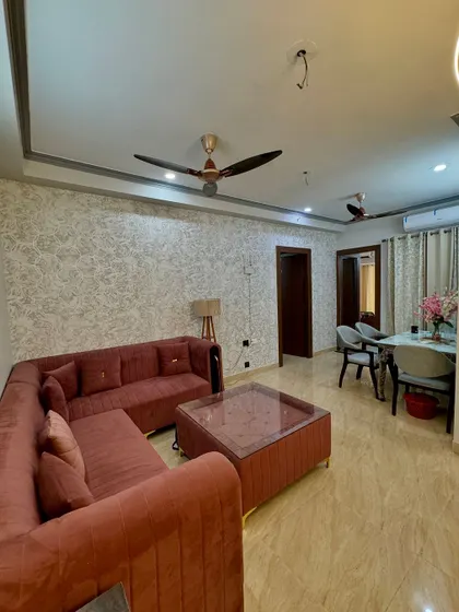 A full view of a living and dining area. The space features a textured wallpaper, a simple false ceiling, and a comfortable L-shaped sofa.