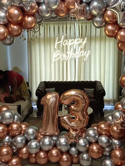 A stylish 13th birthday setup in a living room. This decoration features a large balloon frame made of rose gold and silver chrome balloons, with matching foil balloons for the number '13' and a central neon sign.