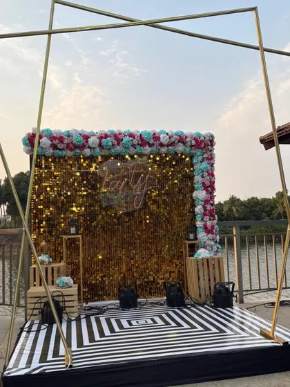 A large-scale outdoor party setup featuring a gold shimmer wall with a 'Let's Party' neon sign, framed by a geometric arch.