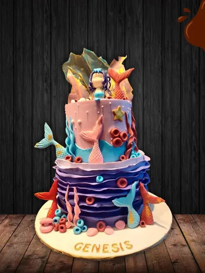 This two-tier mermaid cake is an underwater fantasy. The purple and blue design features multiple mermaid tails, coral, and a beautiful mermaid figure on top, creating a magical ocean scene.