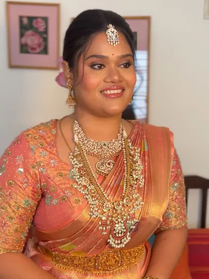 A happy bride showing off her complete look. The unique floral hairstyle perfectly complements her peach-colored silk saree and stunning jewelry.
