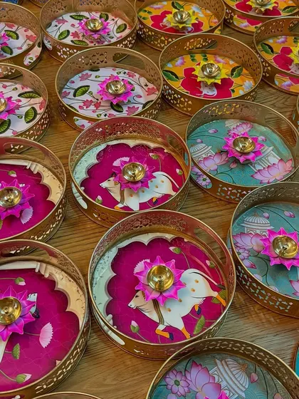 A collection of decorative trays used as part of a bulk order for wedding return favors. Each tray features unique, colorful Indian folk art designs.
