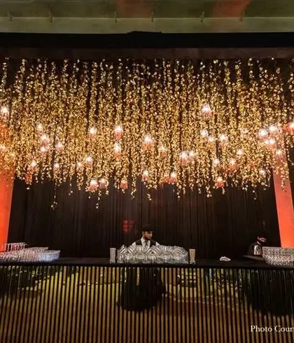 This modern bar counter features a black slatted wood design. The space above is decorated with a stunning installation of hanging flowers and lights, creating a dramatic and sophisticated look for an evening event.
