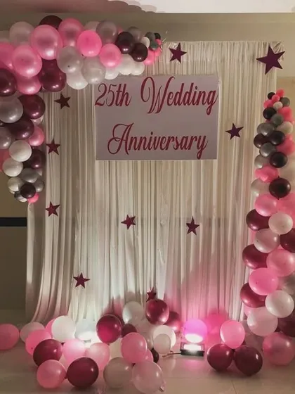 A classic 25th wedding anniversary backdrop with a white curtain and a sign. The setup is framed by a balloon arch in shades of pink, maroon, and white, with star cutouts adding a whimsical touch.