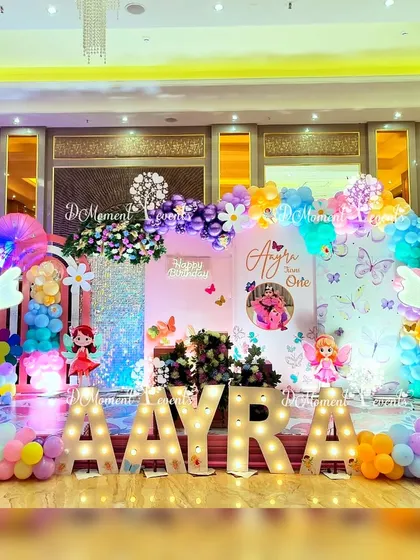 A bright and colorful fairy and butterfly stage setup, featuring a sequin wall, floral arrangements, and light-up marquee letters for a vibrant celebration.