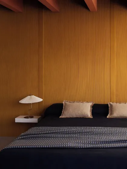 A bedroom featuring the 'Colonne' wallcovering as a headboard. The minimalist lines echo classical temples, while the light reveals delicate textures and refined details, creating a space of simple grandeur.