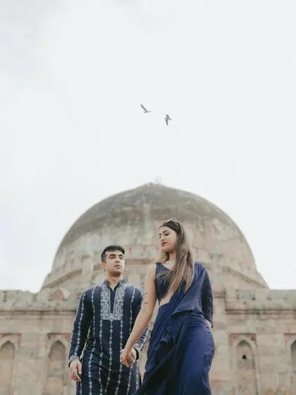A stunning shot of the couple walking hand-in-hand in front of a historic tomb in Delhi. The deep blue of their outfits contrasts beautifully with the ancient monument, while birds in the sky add a touch of serendipity.