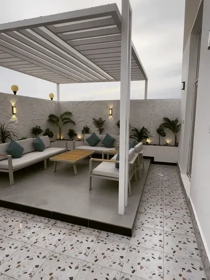 A bright and airy terrace design featuring a white metal pergola over a raised concrete platform. The space is furnished with modern outdoor sofas and planters, set against a textured white wall with accent lighting.
