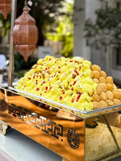A mountain of Dahi Bhalla, beautifully displayed in a golden serving dish. The soft lentil dumplings are soaked in creamy yogurt and garnished with pomegranate seeds and nuts.