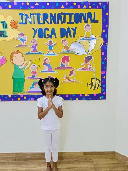 A proud little yogi in her white yoga outfit, standing in a prayer pose in front of our International Yoga Day display board.