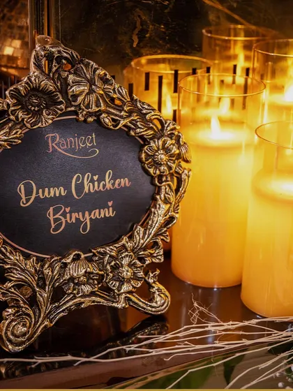 An ornate, framed sign for our Dum Chicken Biryani, set against a backdrop of warm candlelight. This dish is a cornerstone of our traditional Indian menu, slow-cooked to perfection.