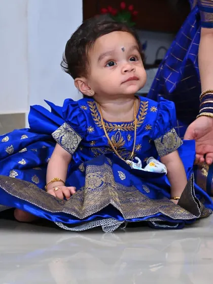 This image details the fine craftsmanship of a traditional Pattu Pavadai. The royal blue silk top features puffed sleeves with zari borders and delicate neckline embroidery, designed to be comfortable against a baby's skin.