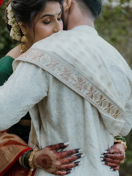 A close-up shot focusing on the details of a tender hug. The bride's henna-adorned hands and the intricate embroidery on the groom's shawl are beautifully highlighted.
