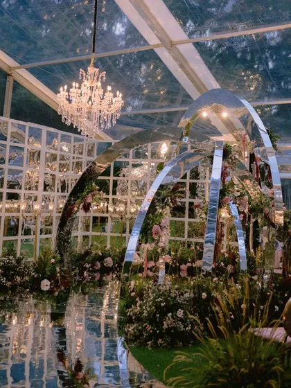 A one-of-a-kind contemporary mandap with mirrored arches, set within a massive glasshouse. This installation, surrounded by dreamy fresh florals, was designed to reflect the couple's modern personality.
