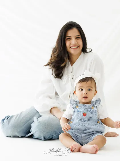 A playful and casual mother-daughter portrait. The baby sits happily in front of her smiling mother, creating a relaxed and authentic family photo.