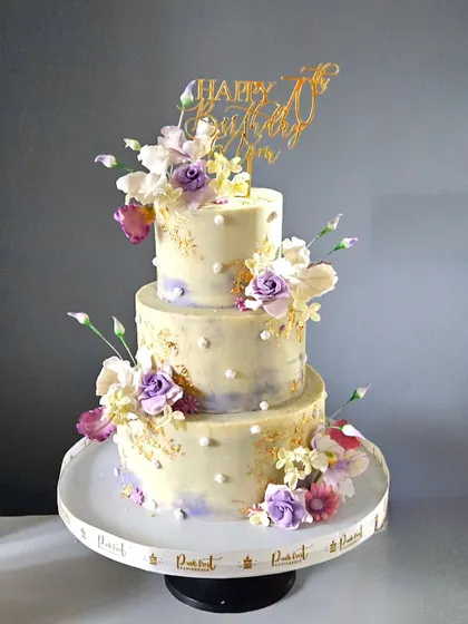 An elegant three-tiered cake to celebrate a 70th birthday. The cake has a subtle lavender and ivory watercolor finish, accented with gold leaf and beautiful, handcrafted sugar flowers.