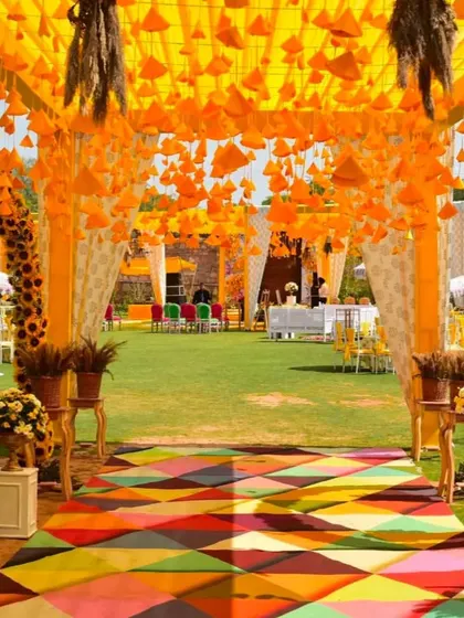 A colorful and festive entrance for a Haldi carnival. The walkway is a vibrant multi-color carpet, leading into a space decorated with hanging orange cones and marigold arches.