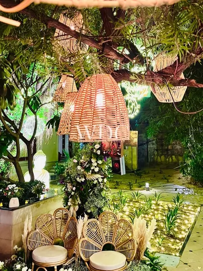 A whimsical seating area in the garden, with peacock chairs set under a tree adorned with hanging cane lanterns.