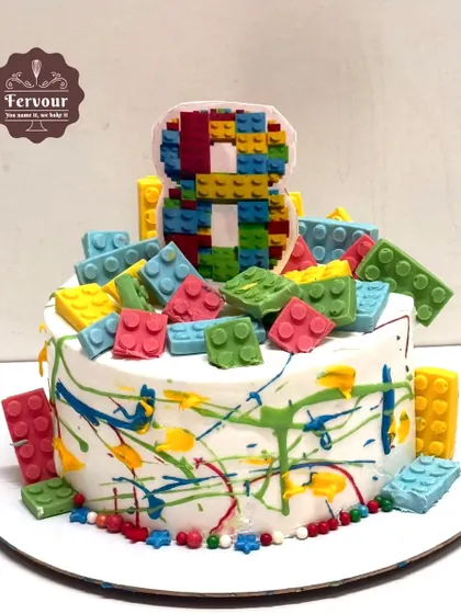 A colorful Lego-themed cake with edible Lego blocks spilling over the top. The paint-splatter effect on the sides adds a fun, creative touch for an 8th birthday.