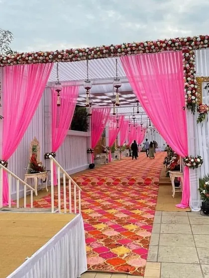A beautiful daytime wedding entrance with pink draping and framed floral arrangements. The colorful patterned carpet adds a festive touch to the elegant white structure.