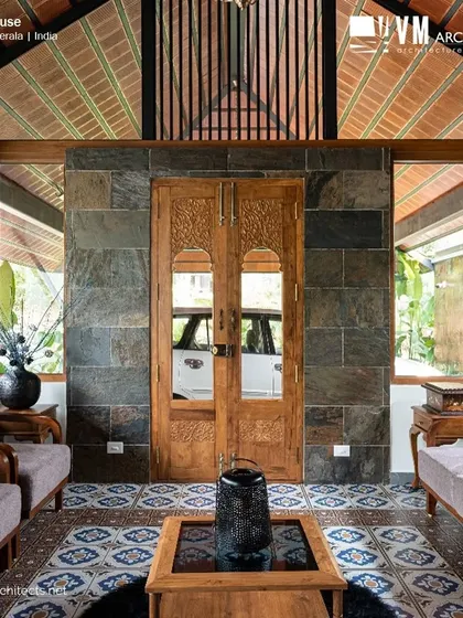 The traditional sitting room at Kuttiyil House, with its patterned floor, stone accent wall, and carved wooden doors, creates a space rich in heritage and craftsmanship.