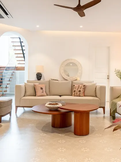 A bright and airy living room in a Goa villa, designed to feel fresh and welcoming. The light-colored sofas and armchairs are paired with natural wood tables, and the patterned floor tiles add a touch of local character. The open archway provides a glimpse of the staircase, enhancing the sense of flow.